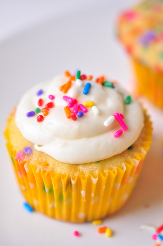 A rainbow sprinkle filled cupcake with cream cheese frosting topped with more rainbow sprinkles. 