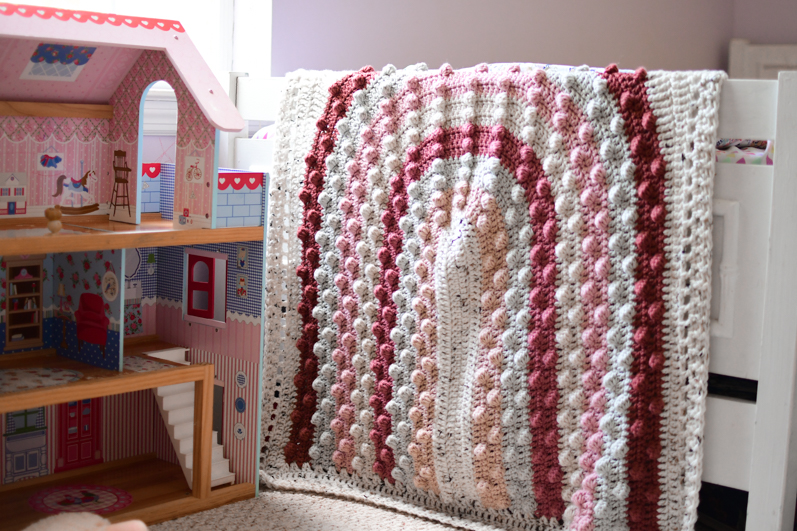 A rainbow bobble blanket draped over the edge of a bed in a child's room. 