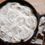 A pan of sourdough cinnamon rolls with cream cheese icing, baked in a cast iron skillet and cooling on a wooden cutting board.