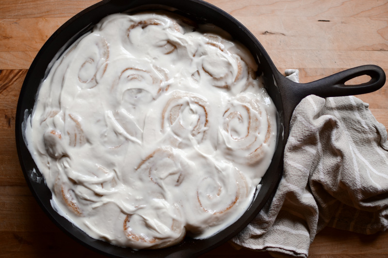 A pan of sourdough cinnamon rolls with cream cheese icing, baked in a cast iron skillet and cooling on a wooden cutting board. 