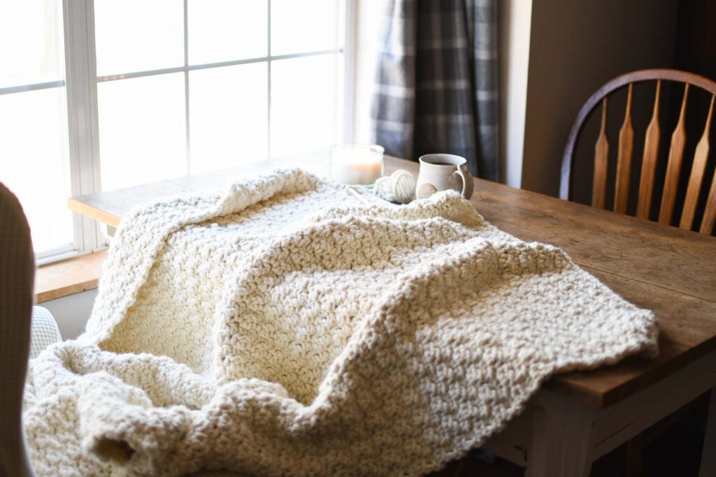 A chunky crochet blanket laying across a table with a candle, cup of coffee, and yarn and hook. 