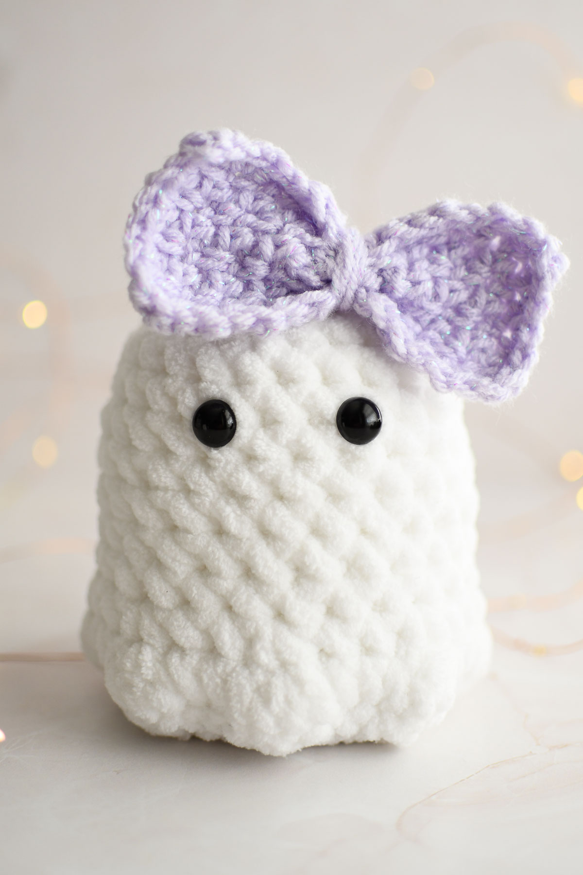 A crochet ghost plushie wearing a large lavender bow. 