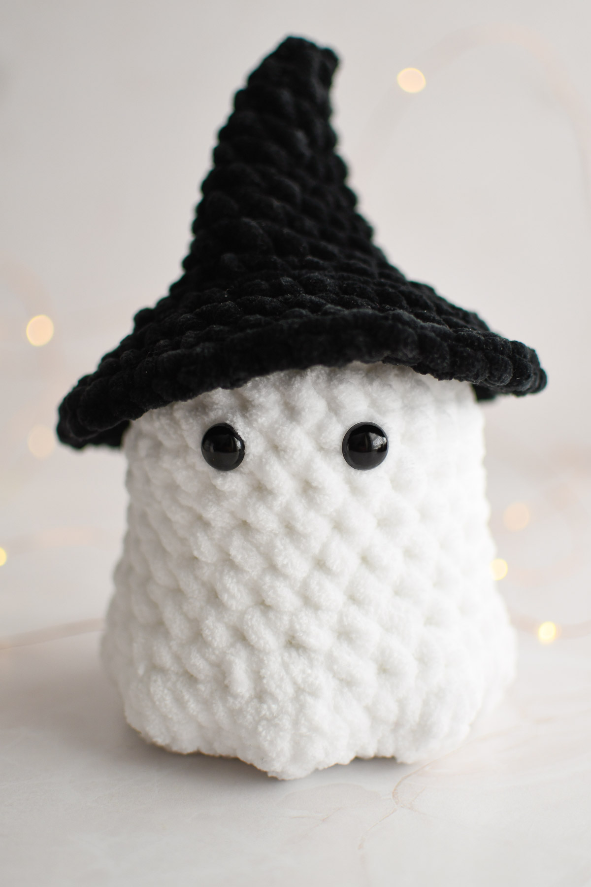 A crochet ghost plushie wearing a large black witch hat. 